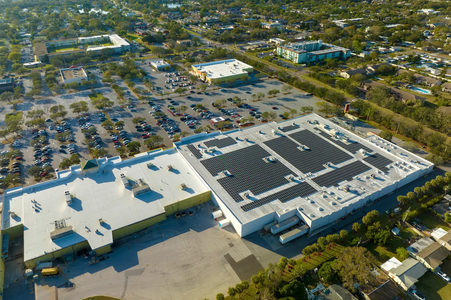 Aerial view of a large commercial building with solar panels installed on the roof and a full parking lot surrounded by trees and residential buildings.