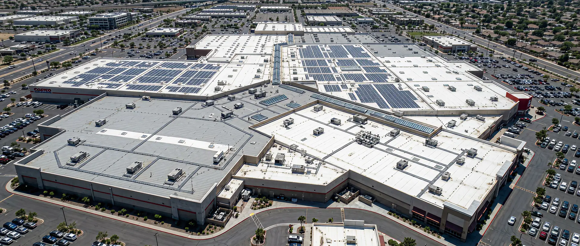 Aerial view of a large retail shopping center with extensive solar panels installed on several white and gray rooftops and surrounding parking lots.