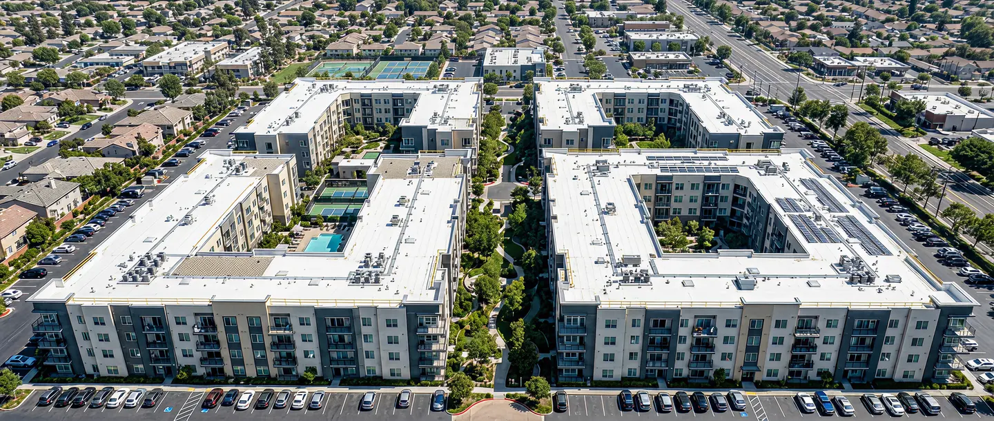 Aerial view of two large, U-shaped multi-family apartment buildings with outdoor pools, tennis courts, parking lots, and green walking paths between them in a suburban neighborhood.