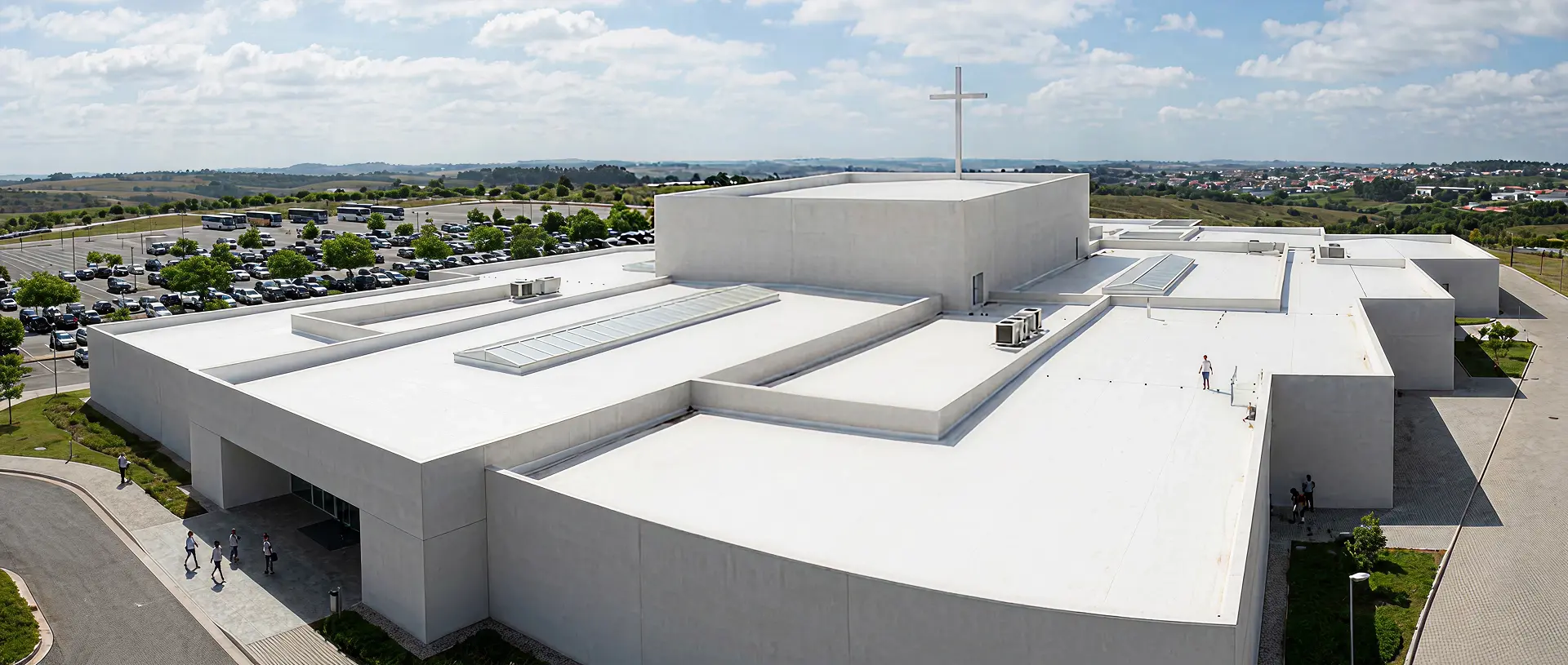 A large, modern white church building with a flat roof and a tall cross on top, surrounded by a parking lot and green landscape under a partly cloudy sky.
