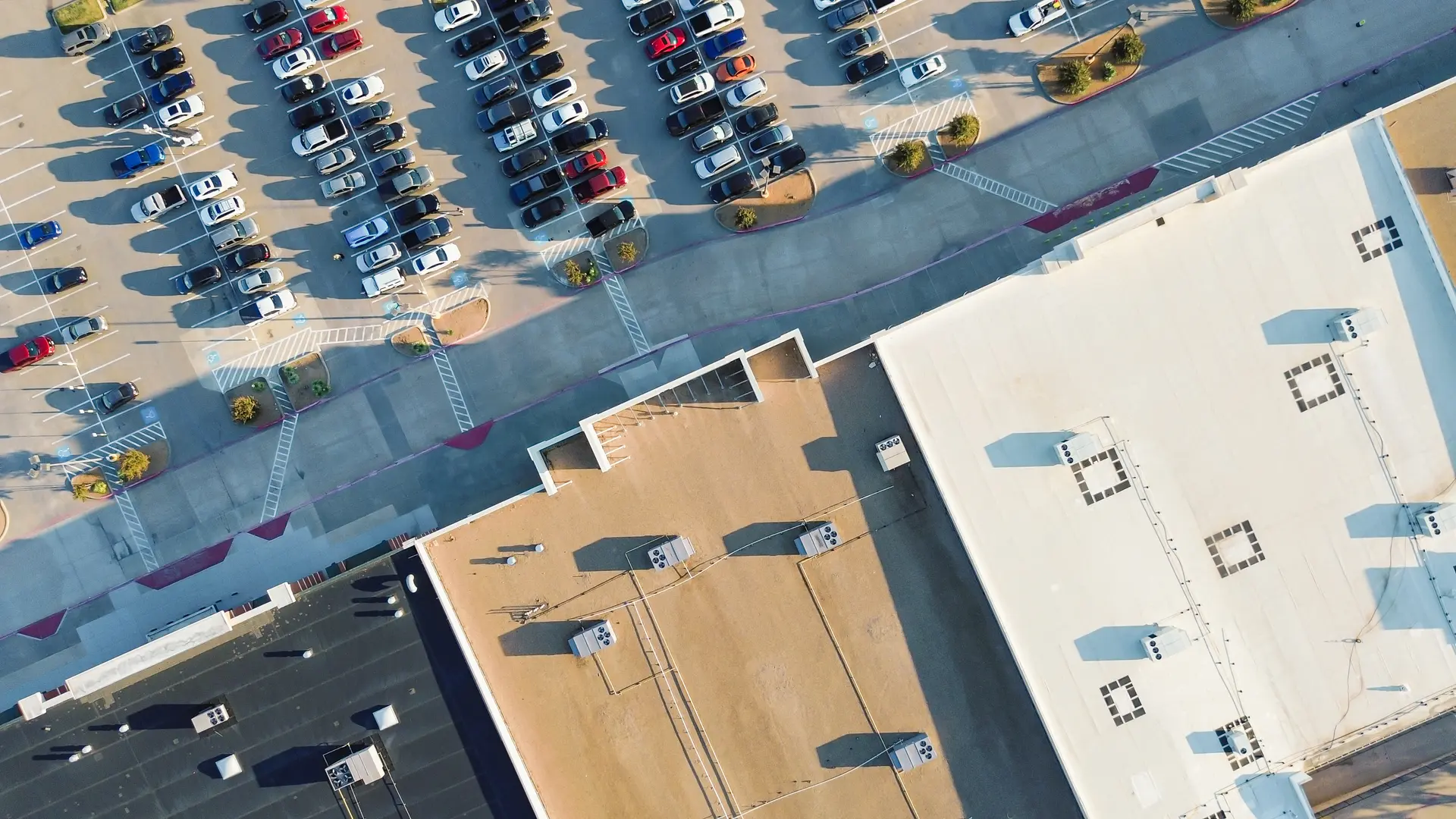 Aerial view of a parking lot filled with cars next to buildings with flat roofs and visible HVAC units.