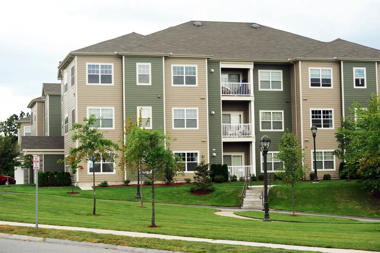 Three-story apartment building with beige and green siding, white balconies, surrounded by green lawn and small trees.