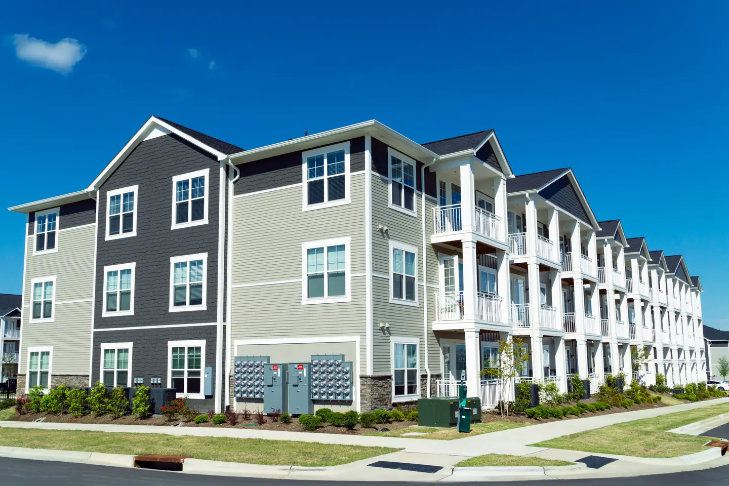Modern three-story apartment building with beige and dark gray siding, white balconies, and large windows under a clear blue sky.