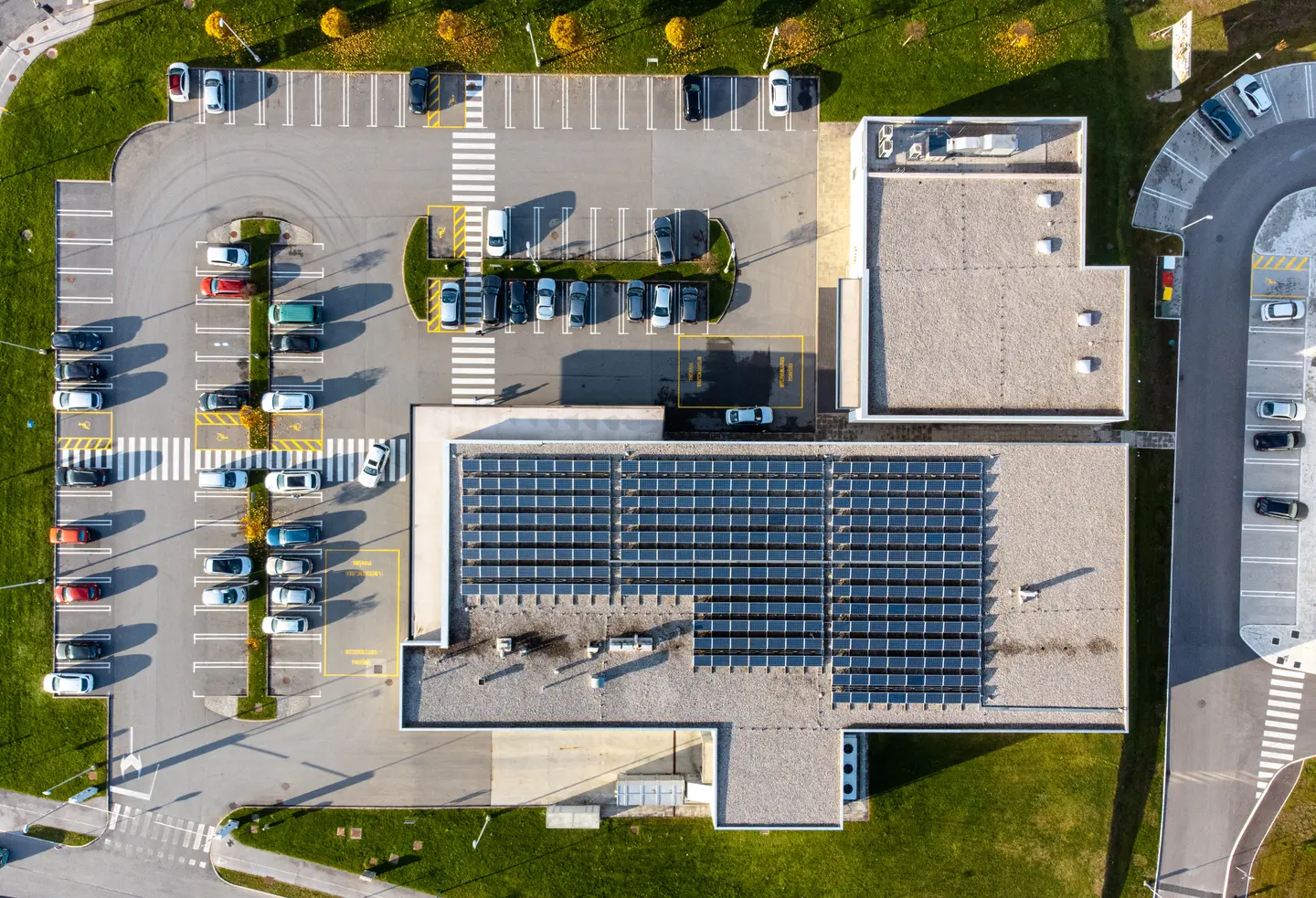 Aerial view of a building with solar panels on the roof and surrounding parking lot with parked cars and green grass.
