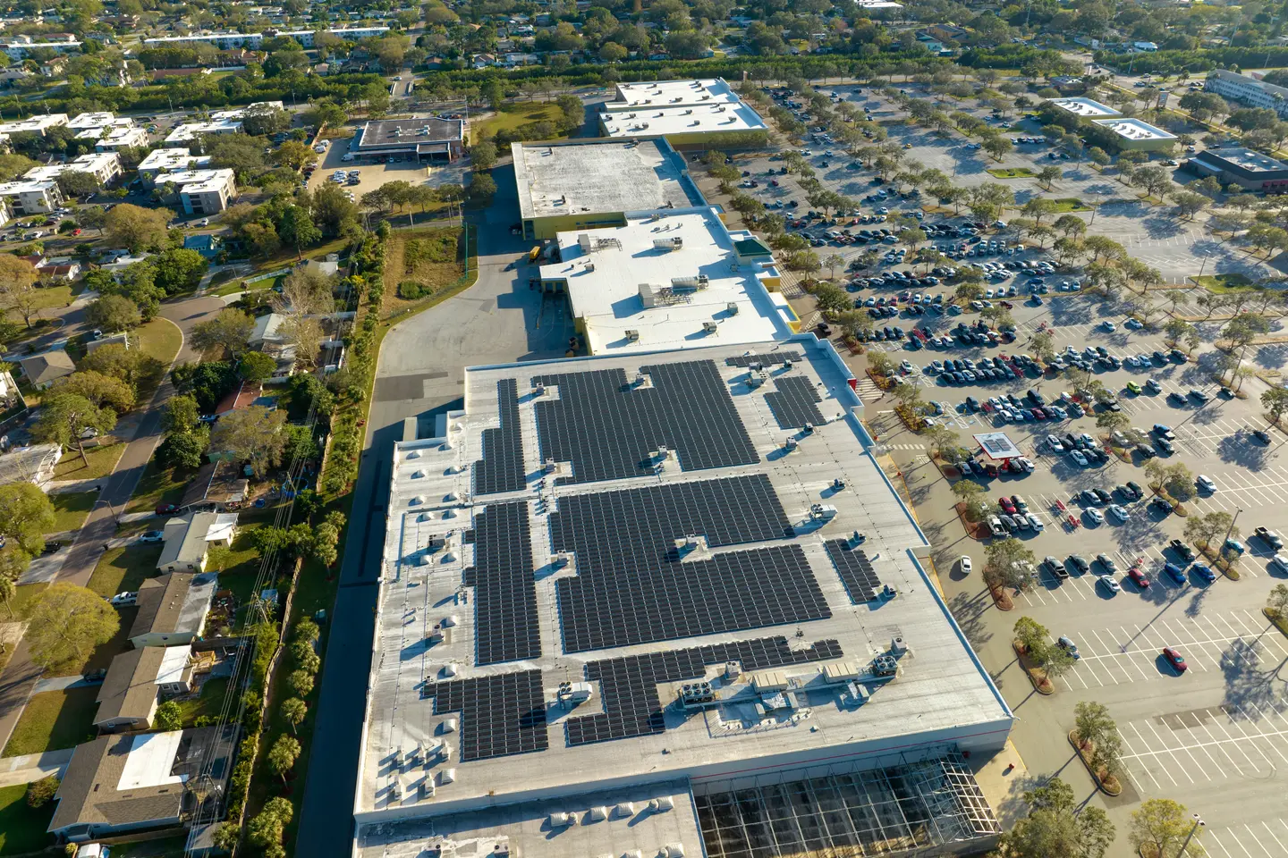 Aerial view of a large building with solar panels installed on its flat roof next to a parking lot with numerous cars and surrounding residential neighborhood.