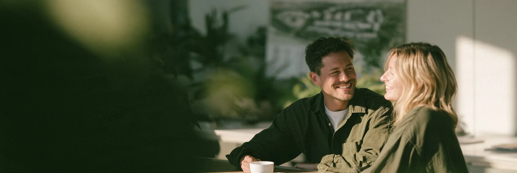 Man and woman laughing over coffee at a table with plants and a pinned site plan behind them