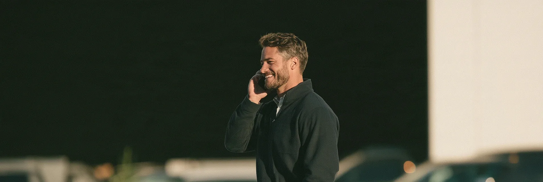 Smiling man in a dark quarter zip talking on his phone outside at golden hour