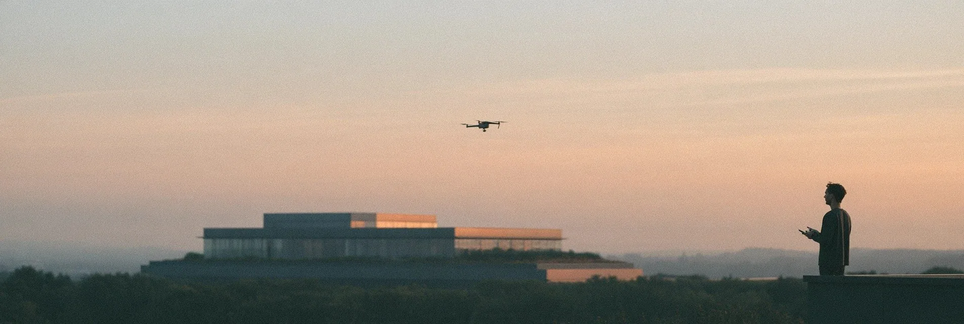 Person flying a drone at dusk over a flat landscape with a low building on the horizon