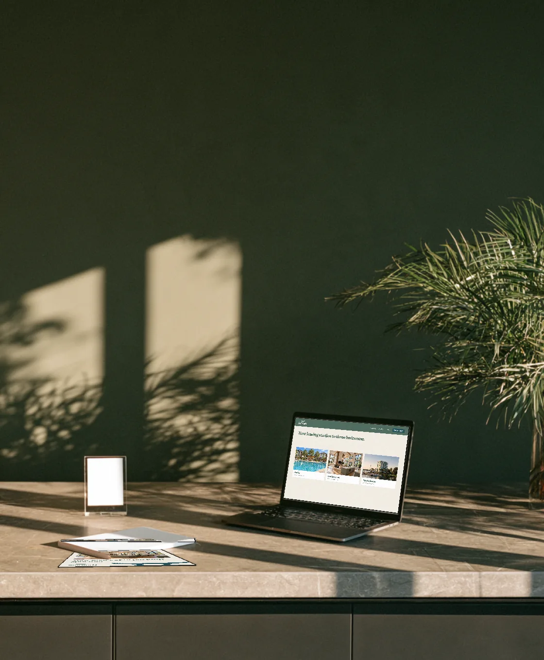 Laptop on a stone counter showing an apartment website, beside a small framed card and printed pages, with palm shadows on the wall