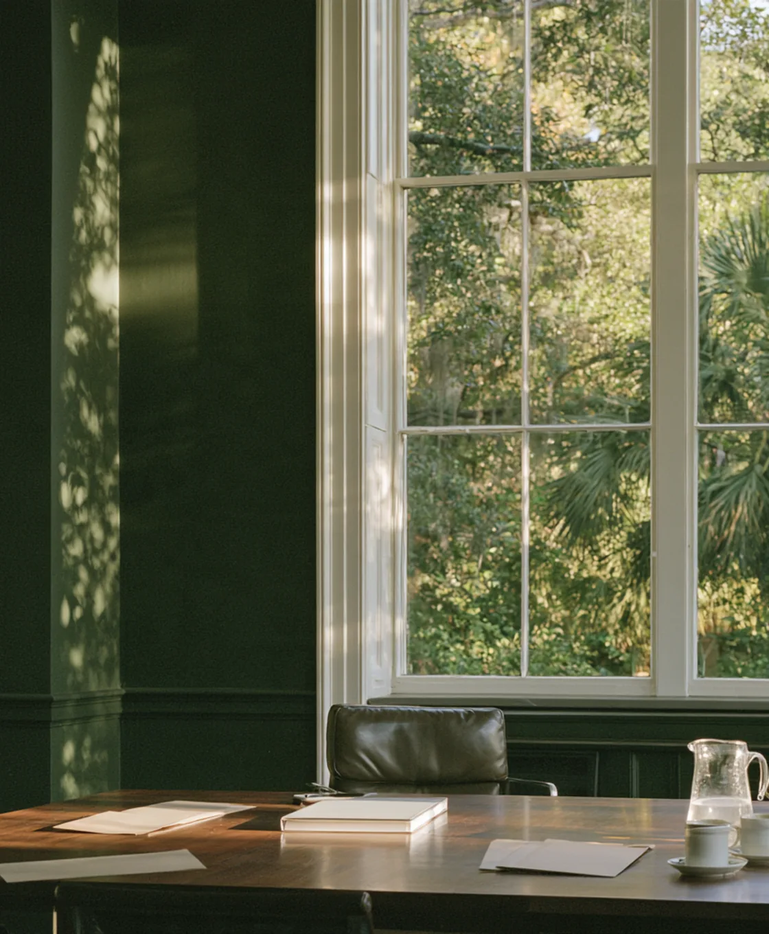 Empty meeting room with a wooden table, papers and coffee cups in front of a tall window onto green trees