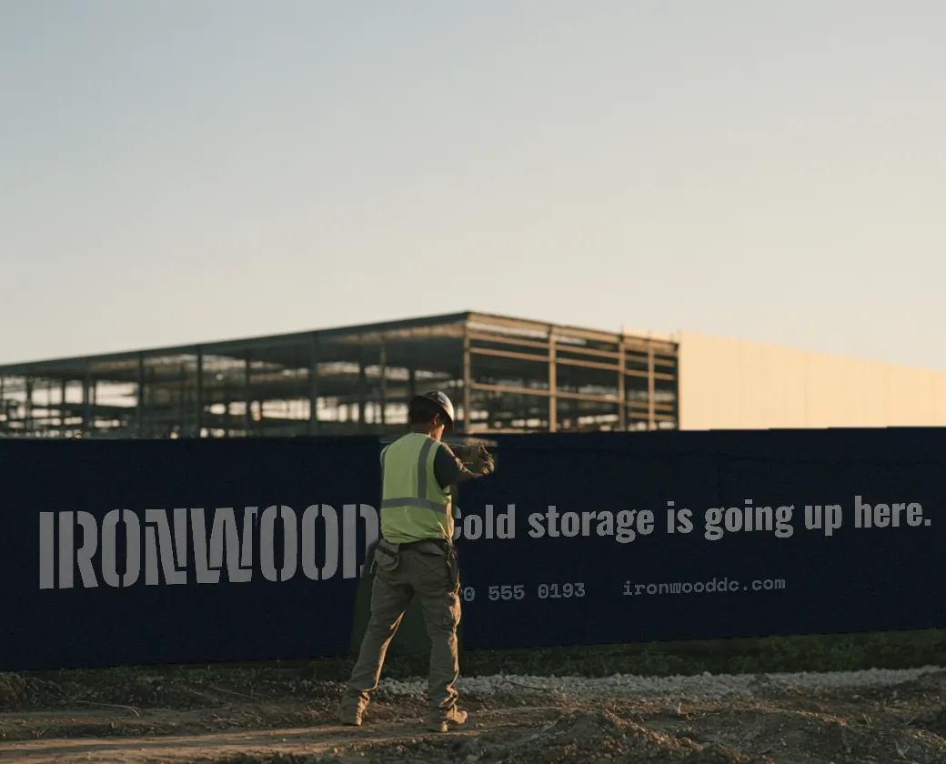 Worker in a hard hat and safety vest beside an Ironwood fence wrap reading Cold storage is going up here, with a steel framed building behind
