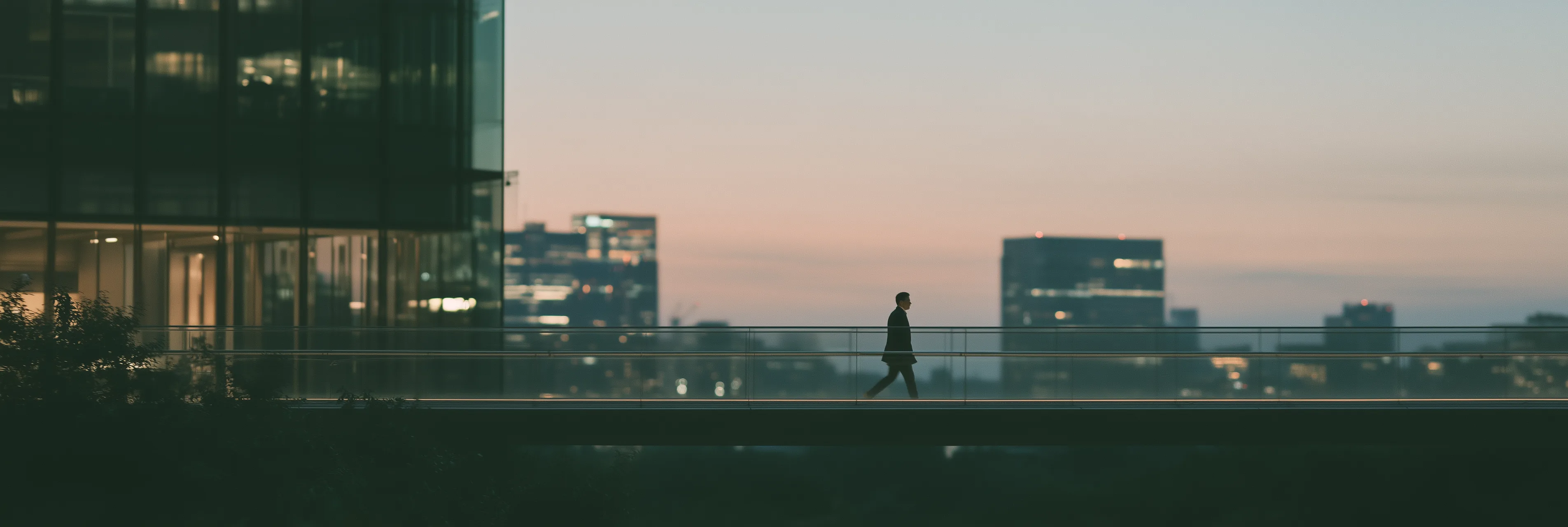 A lone figure crossing a glass skybridge between office towers at dusk, with the city skyline beyond
