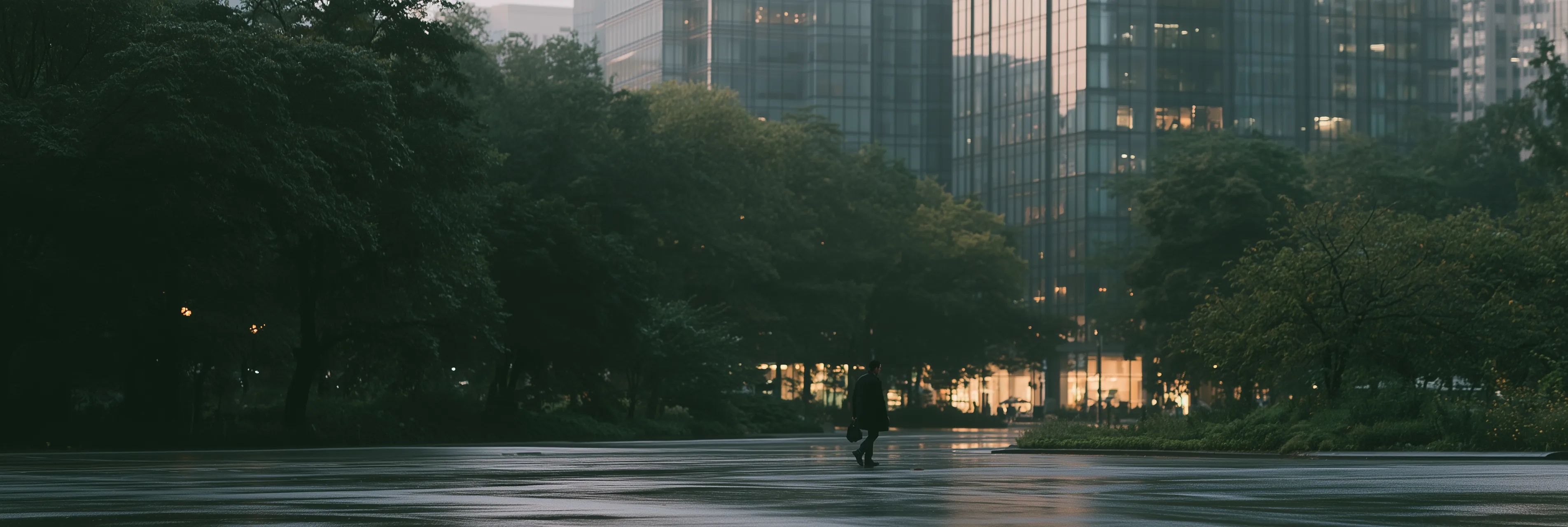 A lone figure crossing a wet city intersection at blue hour, office towers and trees beyond