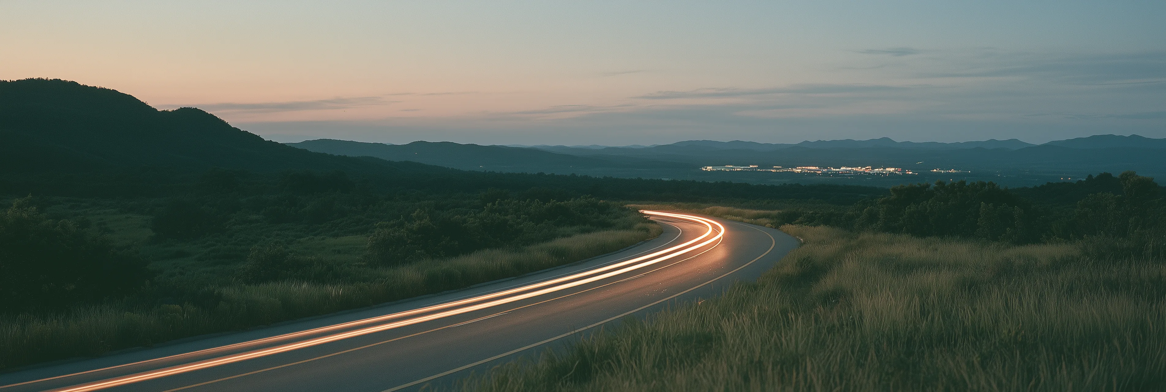 Headlight trails curving along a quiet highway at dusk toward a distant lit business park