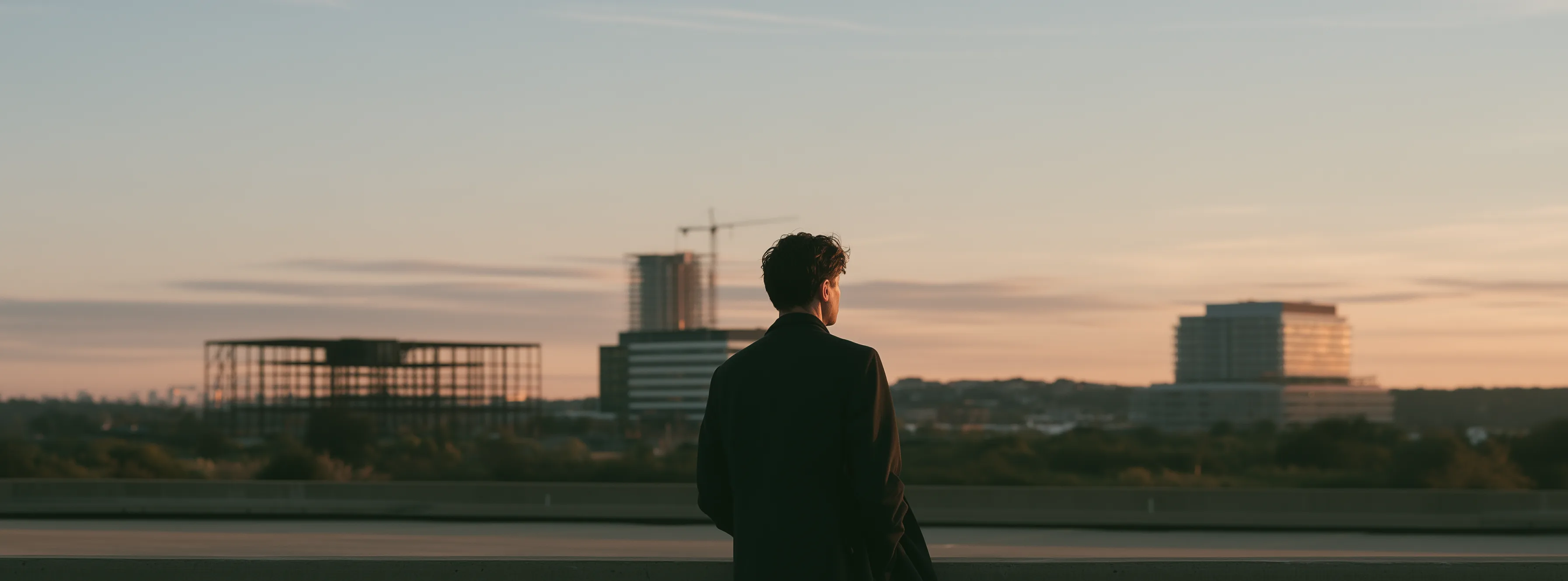 Developer seen from behind at dusk looking out over three projects rising at once: a warehouse frame, an office tower with a crane and a mid rise