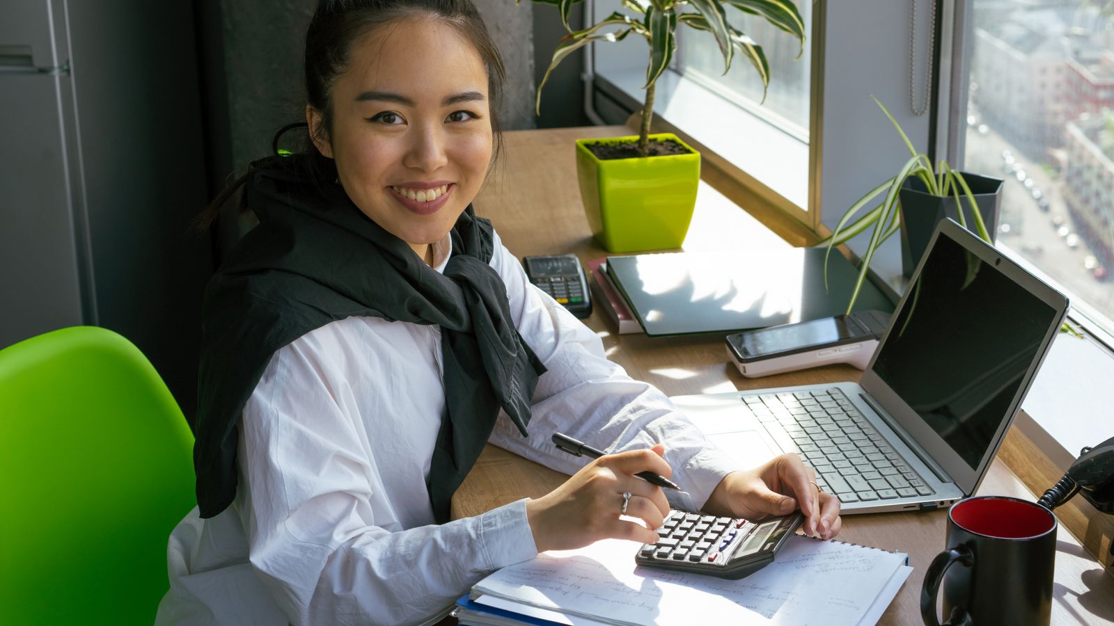 Jeune entrepreneuse souriante calculant la rentabilité de sa boutique en ligne avec une calculatrice, un carnet et un terminal de paiement