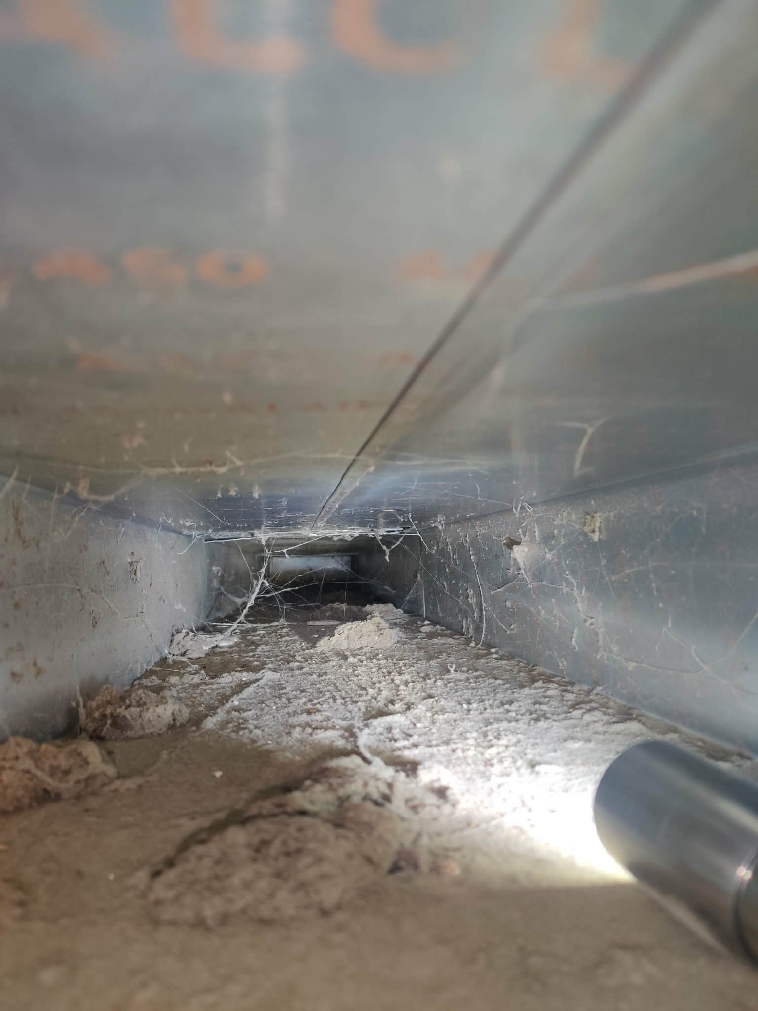 Close-up view inside a dirty, dusty metal duct with cobwebs and a flashlight illuminating the interior.