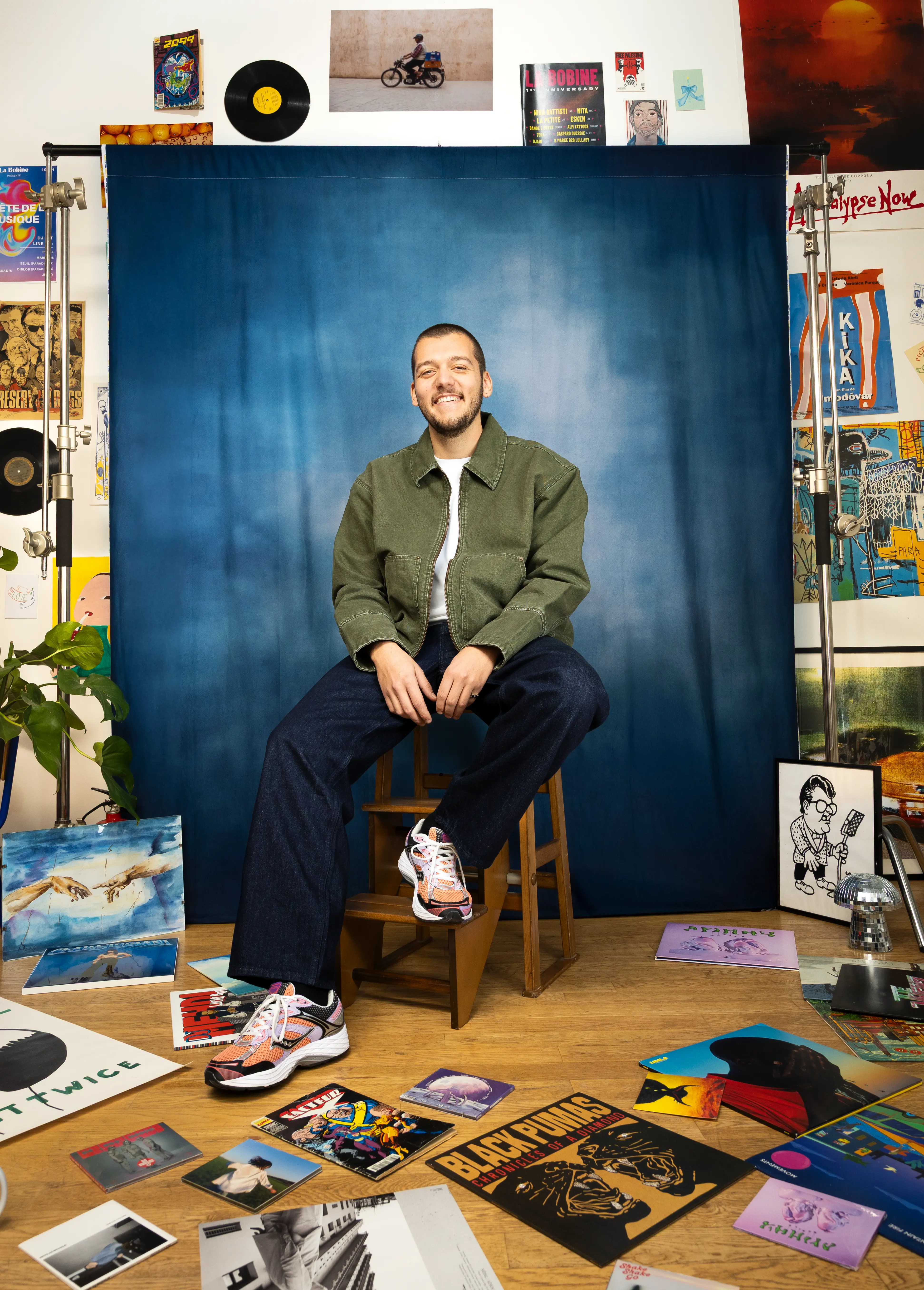 Portrait souriant d'un jeune homme sur un escabeau devant un fond bleu, mettant en avant les artistes émergents.