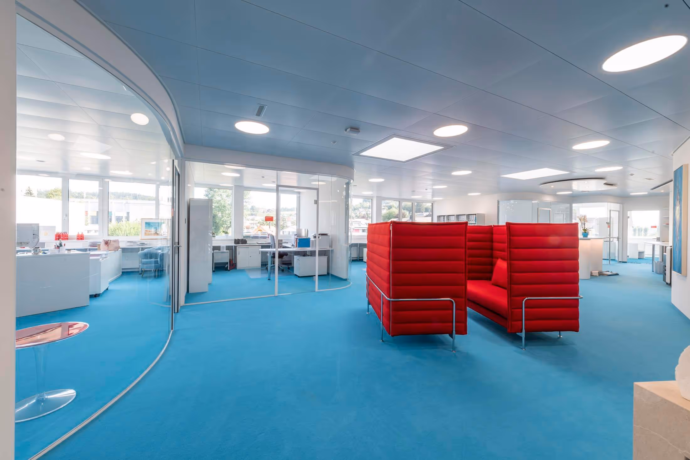 Modern office interior with blue carpet, large windows, glass-walled meeting rooms, and bright red cushioned seating pods.