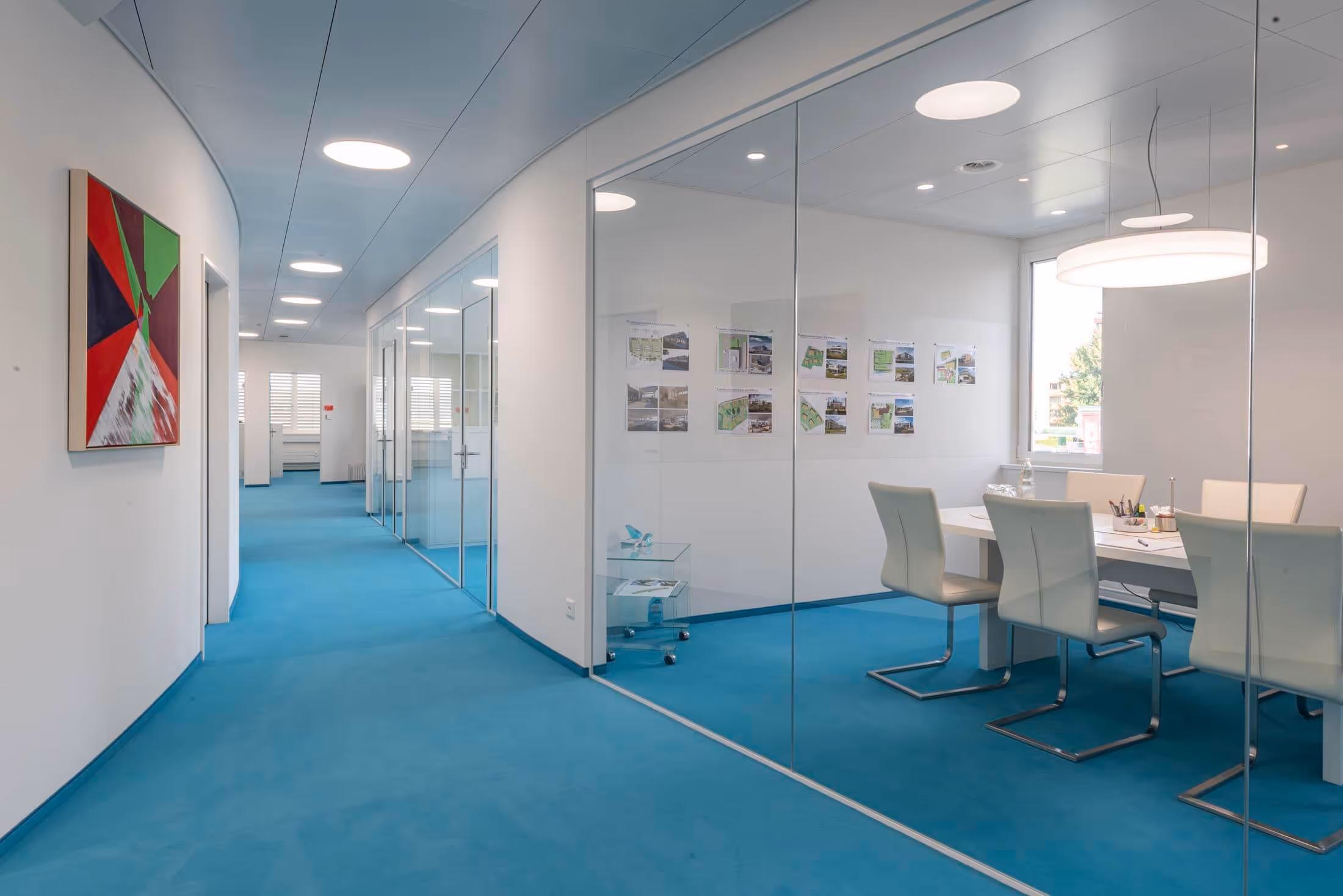 Modern office corridor with blue carpet, glass-walled meeting room with white chairs, and abstract artwork on the wall.