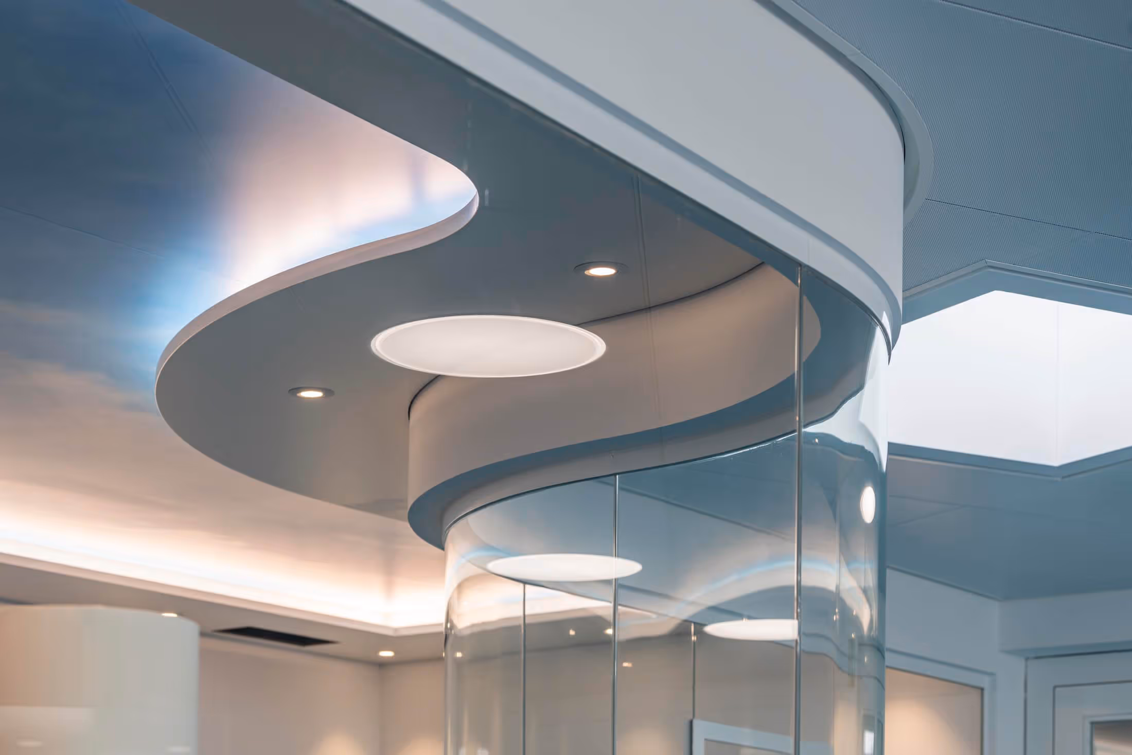 Curved modern ceiling design with circular recessed lighting and a glass wall in a bright interior space.