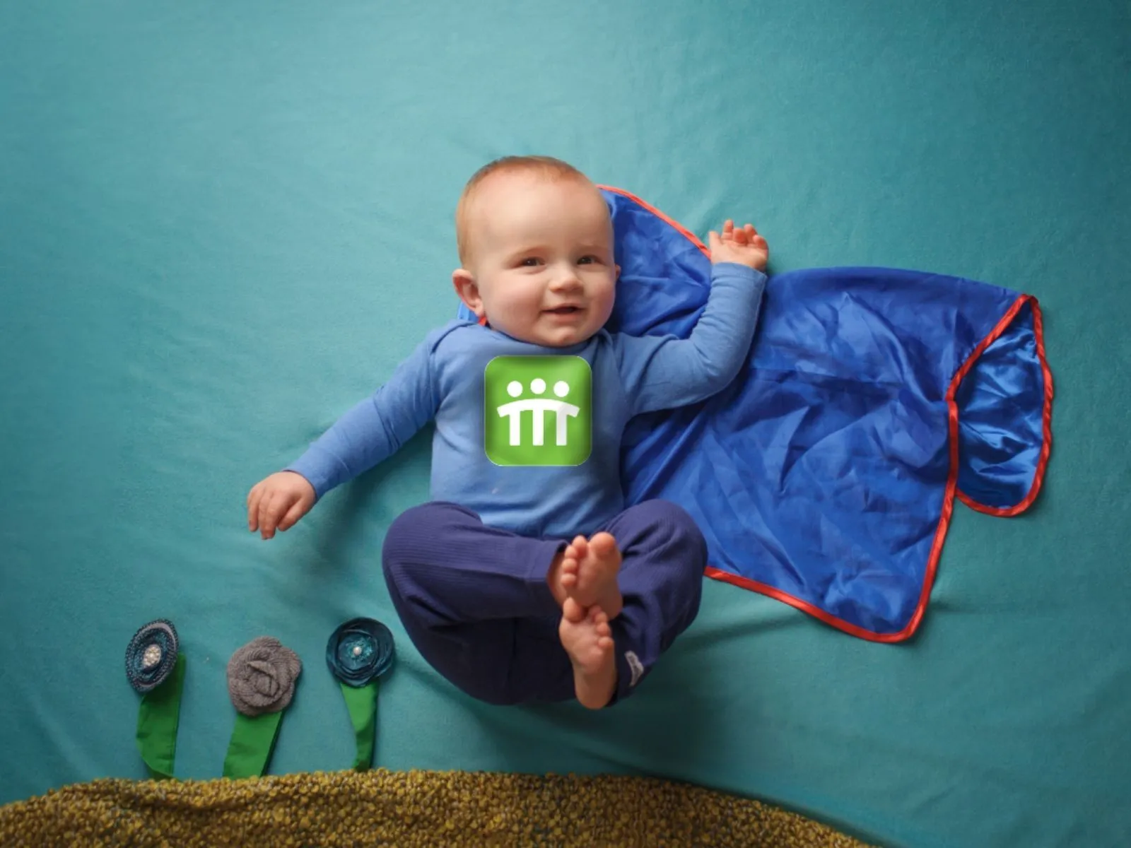A baby boy laying on his back, dressed in a onsie with the MetroEHS logo and a blue superhero cape