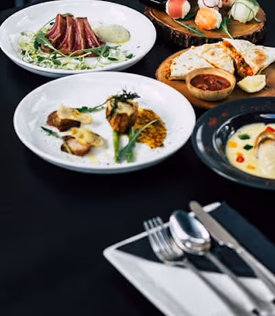 Assortiment de plats gourmets sur table noire comprenant viande tranchée, rouleaux de printemps, quiche avec sauce rouge, et coupe d'ustensiles sur une serviette blanche.