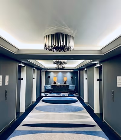 Couloir d'hôtel moderne avec quatre ascenseurs, tapis bleu et blanc, et espace salon avec deux fauteuils bleus et lampe.