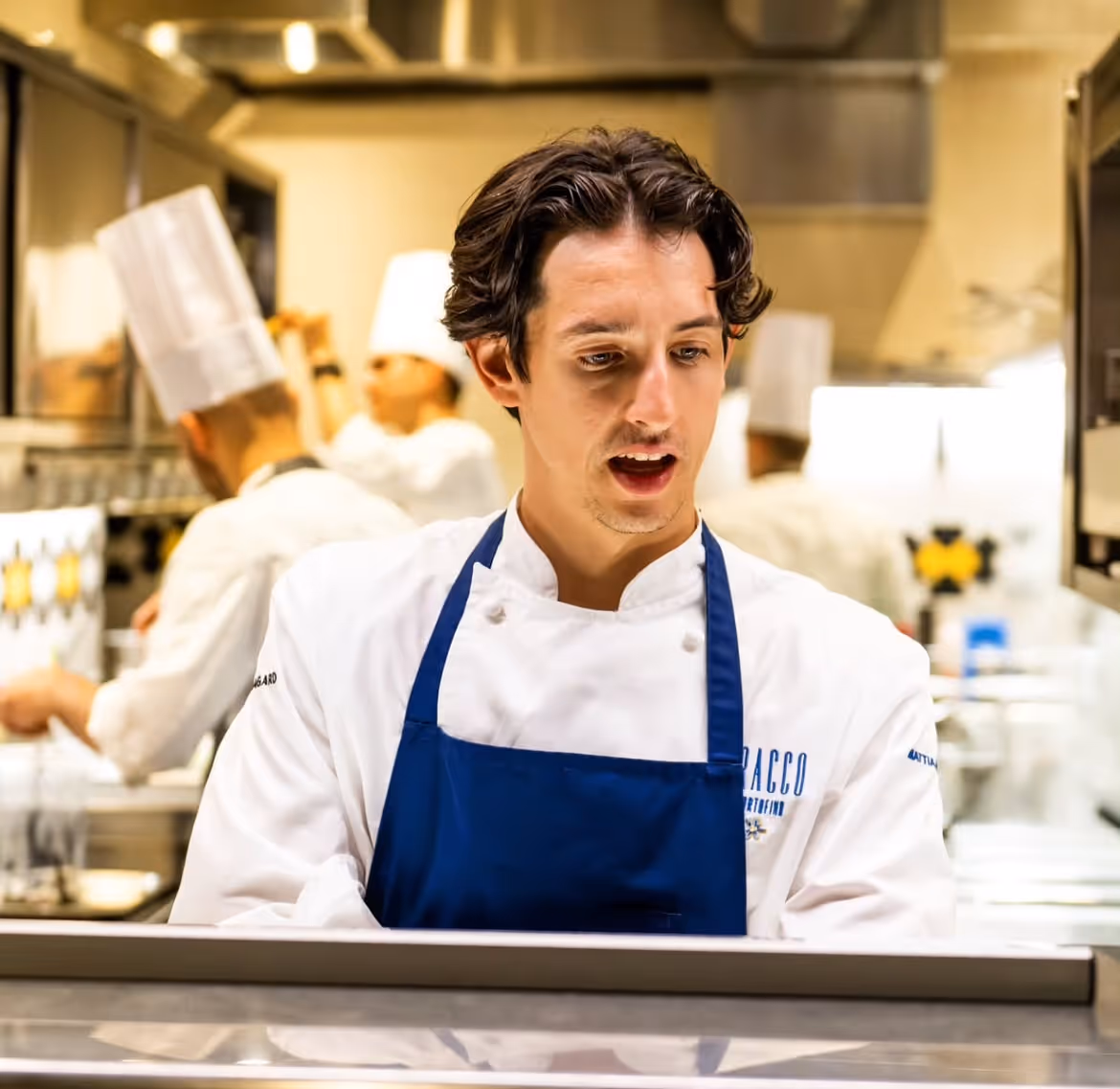 Chef portant une veste blanche et un tablier bleu, travaillant dans une cuisine professionnelle avec des collègues cuisiniers en arrière-plan.