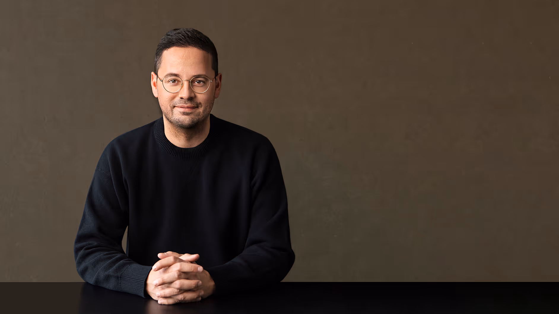Mann mit kurzen Haaren und Brille, der in schwarzem Pullover an einem Tisch sitzt und in die Kamera lächelt.