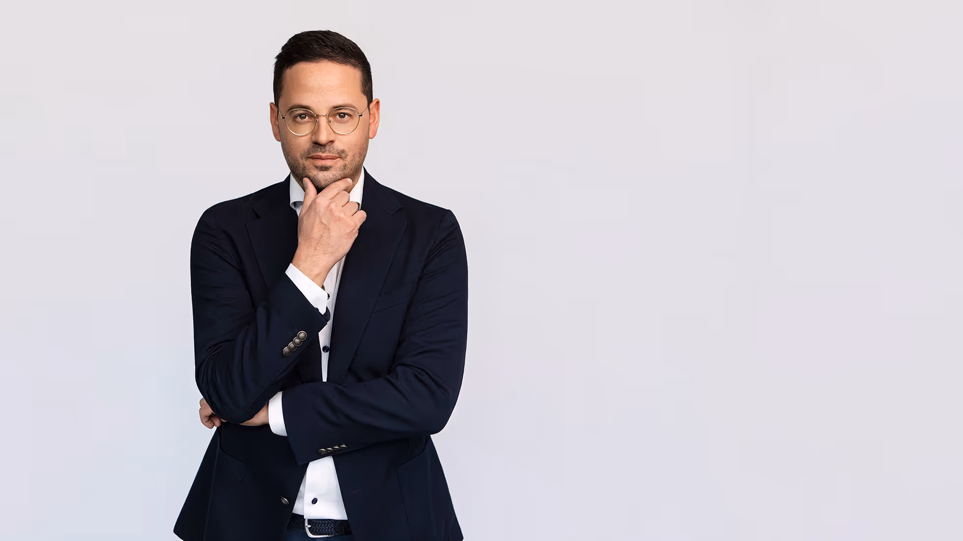 Mann in dunklem Anzug und Brille, der nachdenklich mit der Hand am Kinn steht vor einem hellen Hintergrund.