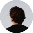 Personne vue de dos avec des cheveux bruns bouclés portant un haut noir sur fond clair.