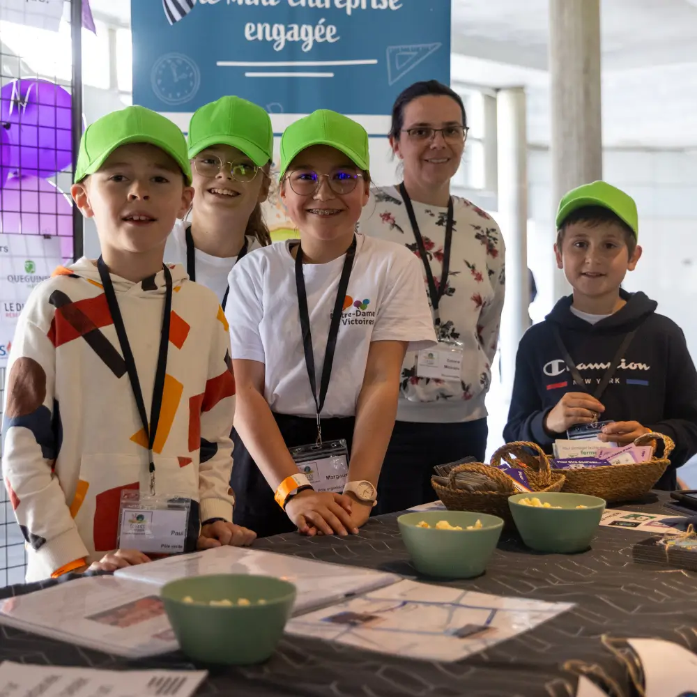 Quatre enfants portant des casquettes vertes et des badges posent derrière une table avec des bols et des paniers lors d'un événement intérieur.