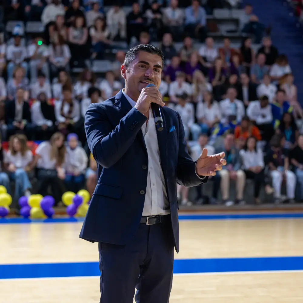 Un homme en costume sombre parlant dans un micro devant un public assis dans un gymnase décoré de ballons jaunes et violets.