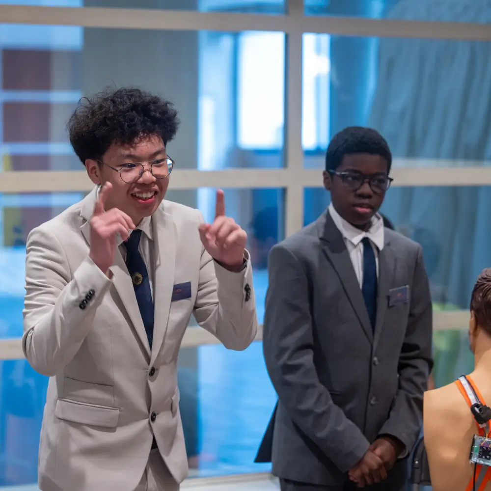 Deux jeunes hommes habillés en costumes formels discutent, l’un gesticulant avec enthousiasme devant une fenêtre.