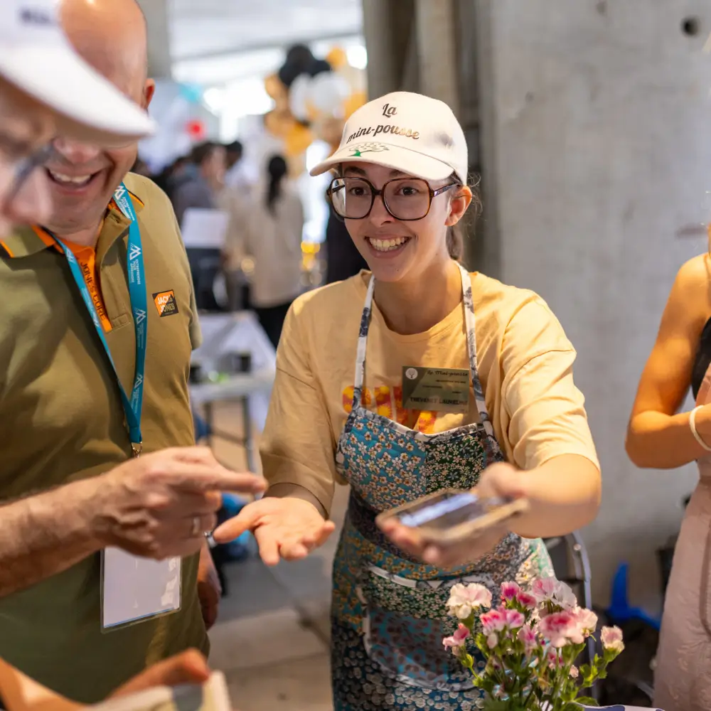 Jeune femme souriante portant une casquette et un tablier, tendant un téléphone portable lors d'un événement animé.