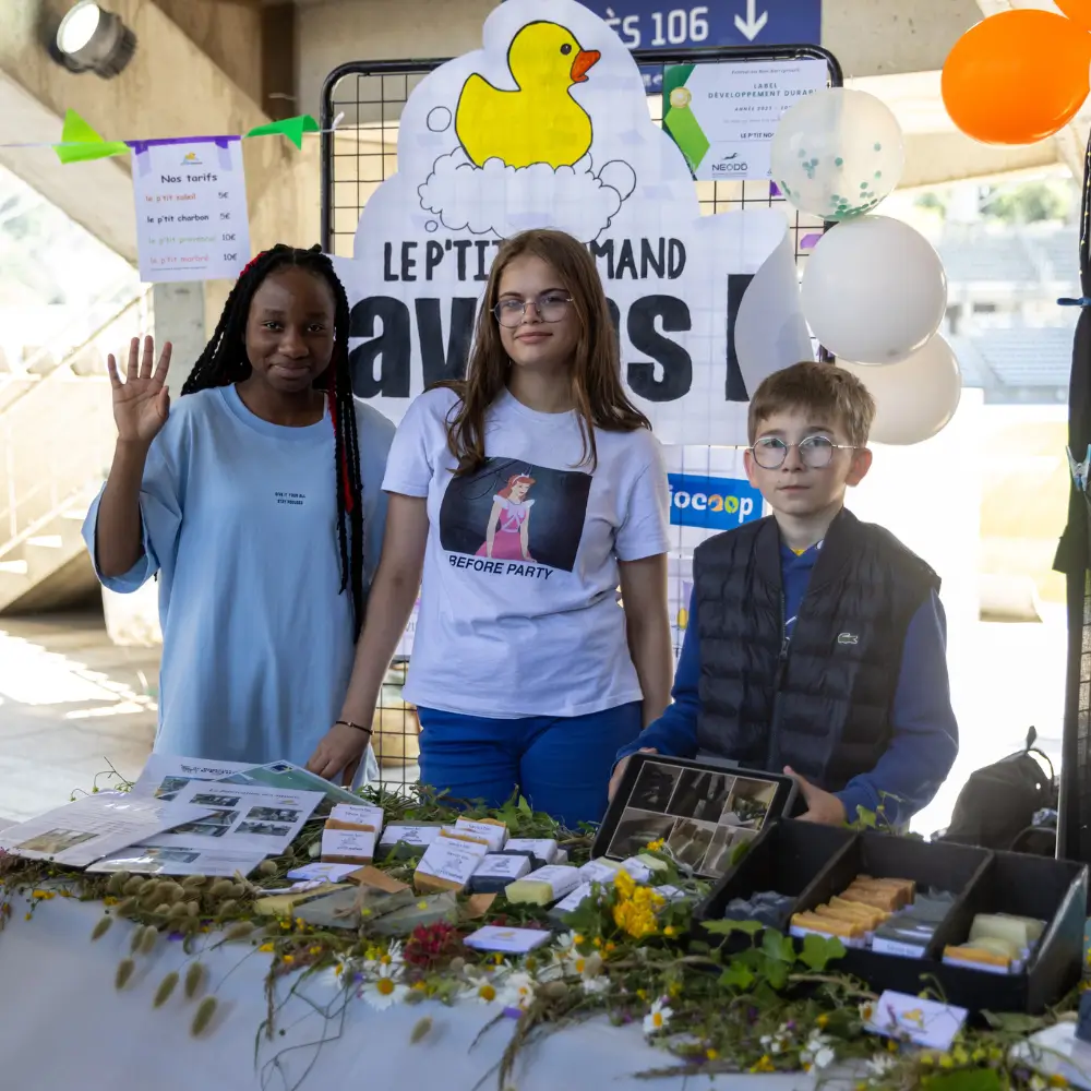 Trois enfants debout derrière un stand décoré avec des savons artisanaux et des fleurs, avec une grande affiche montrant un canard jaune en arrière-plan.