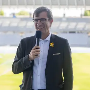 Homme souriant portant des lunettes et un costume sombre tenant un micro dans une arène ou un stade vide.
