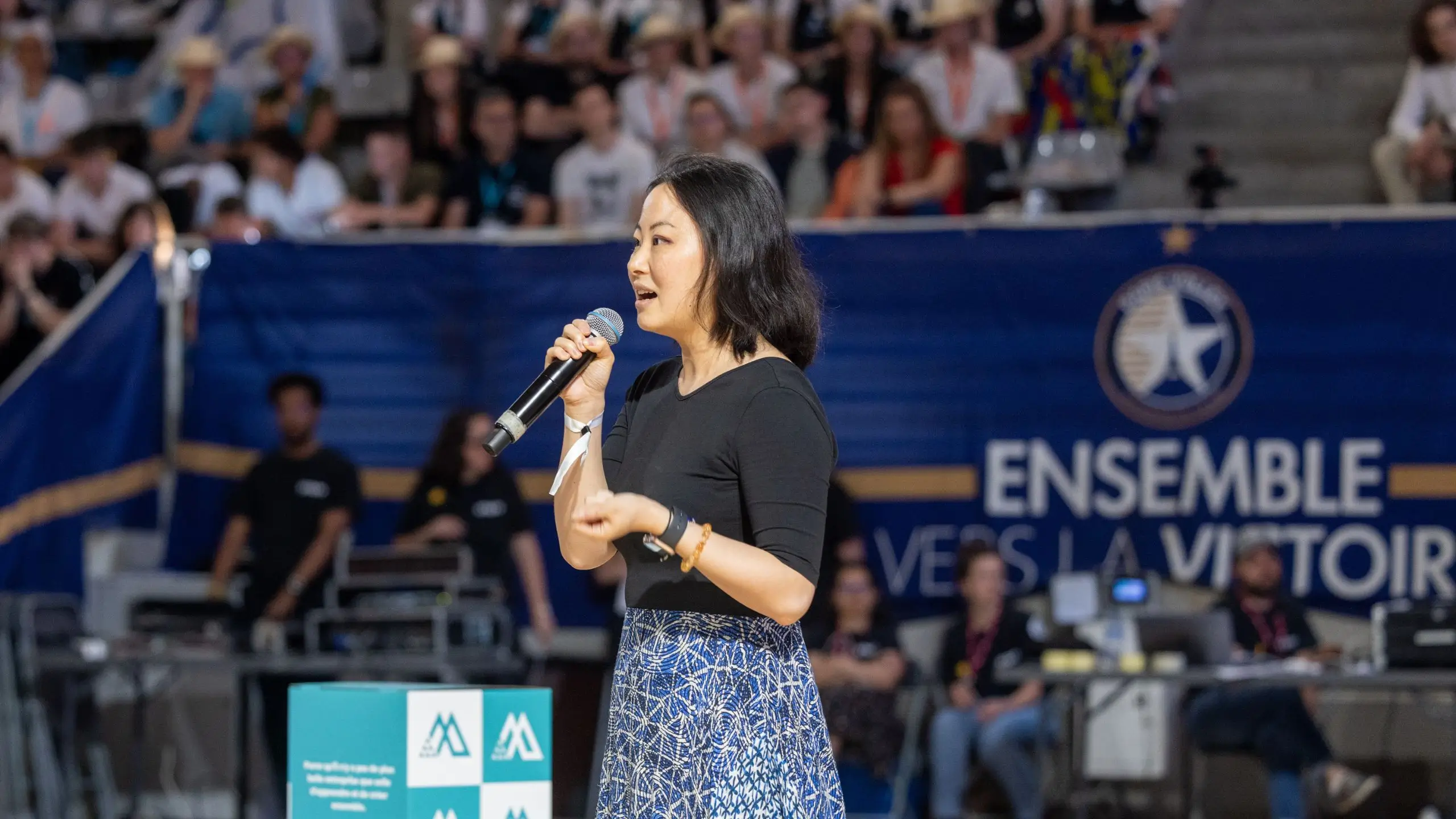 Une femme parlant au micro lors d'un événement devant un public avec une banderole affichant 'ENSEMBLE VERS LA VICTOIRE'.