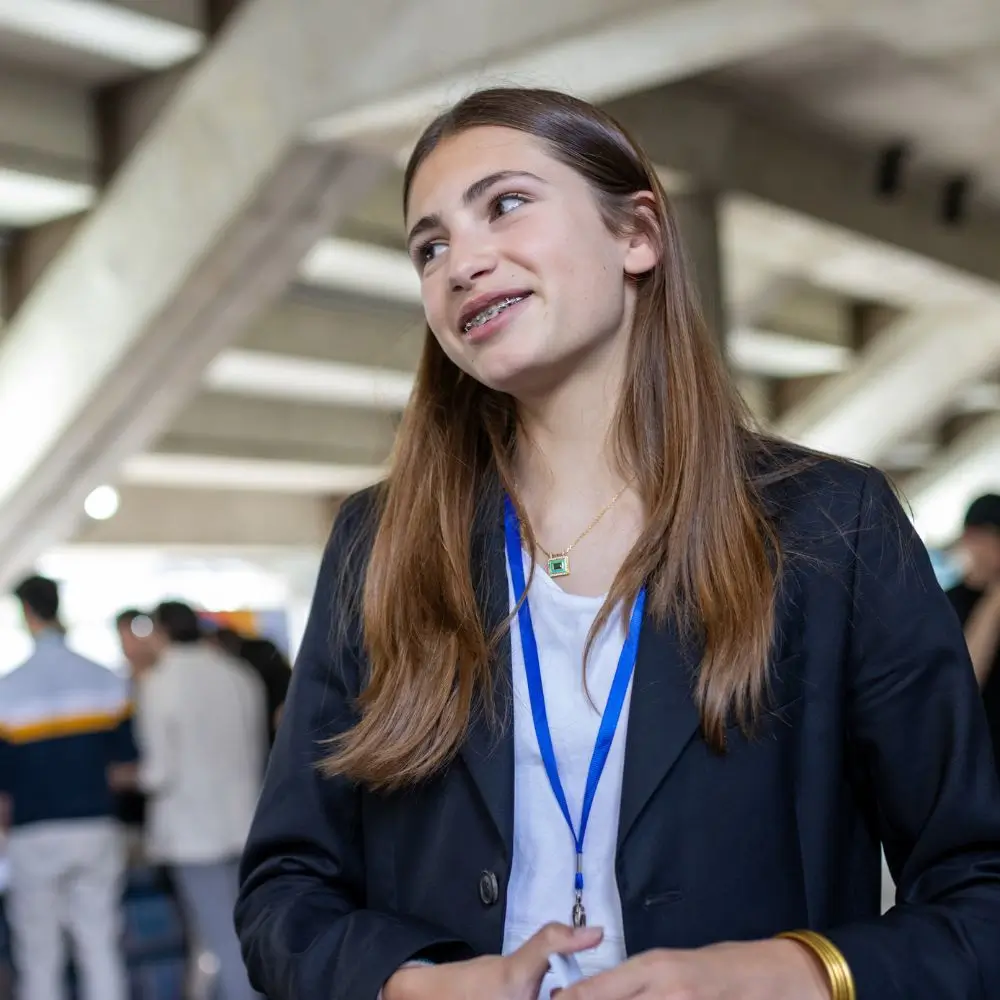 Une jeune femme souriante avec des appareils dentaires, portant un badge bleu et une veste noire dans un espace intérieur animé.