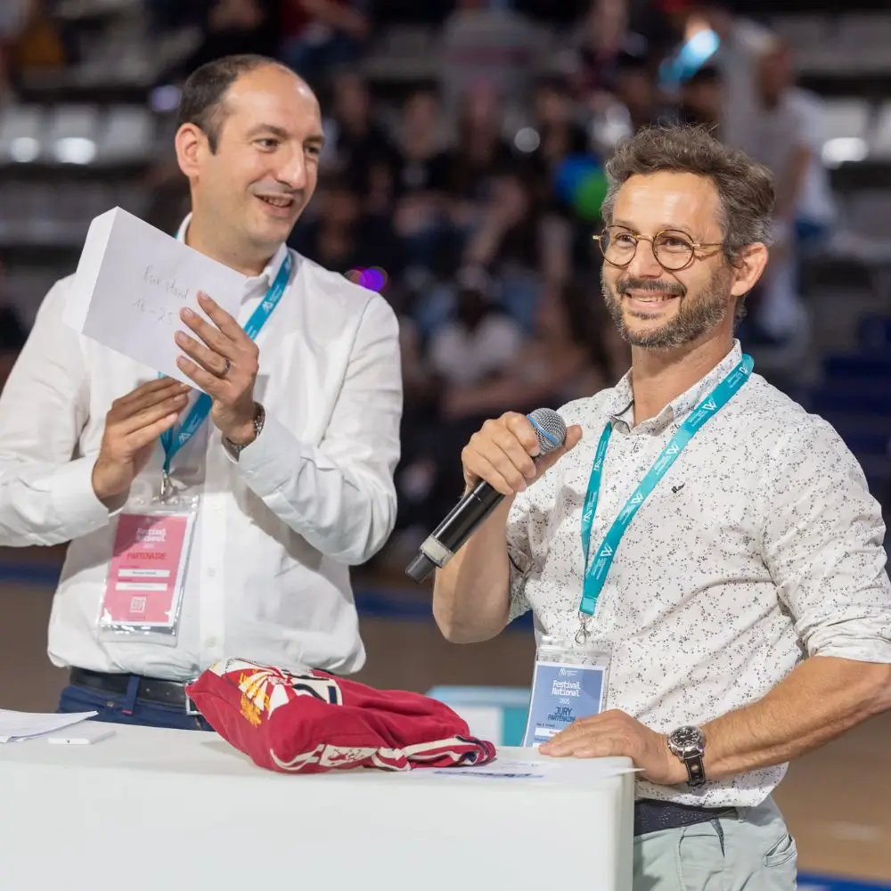 Deux hommes portant des badges de participant discutent à une table, l'un tenant un microphone et l'autre une enveloppe blanche.