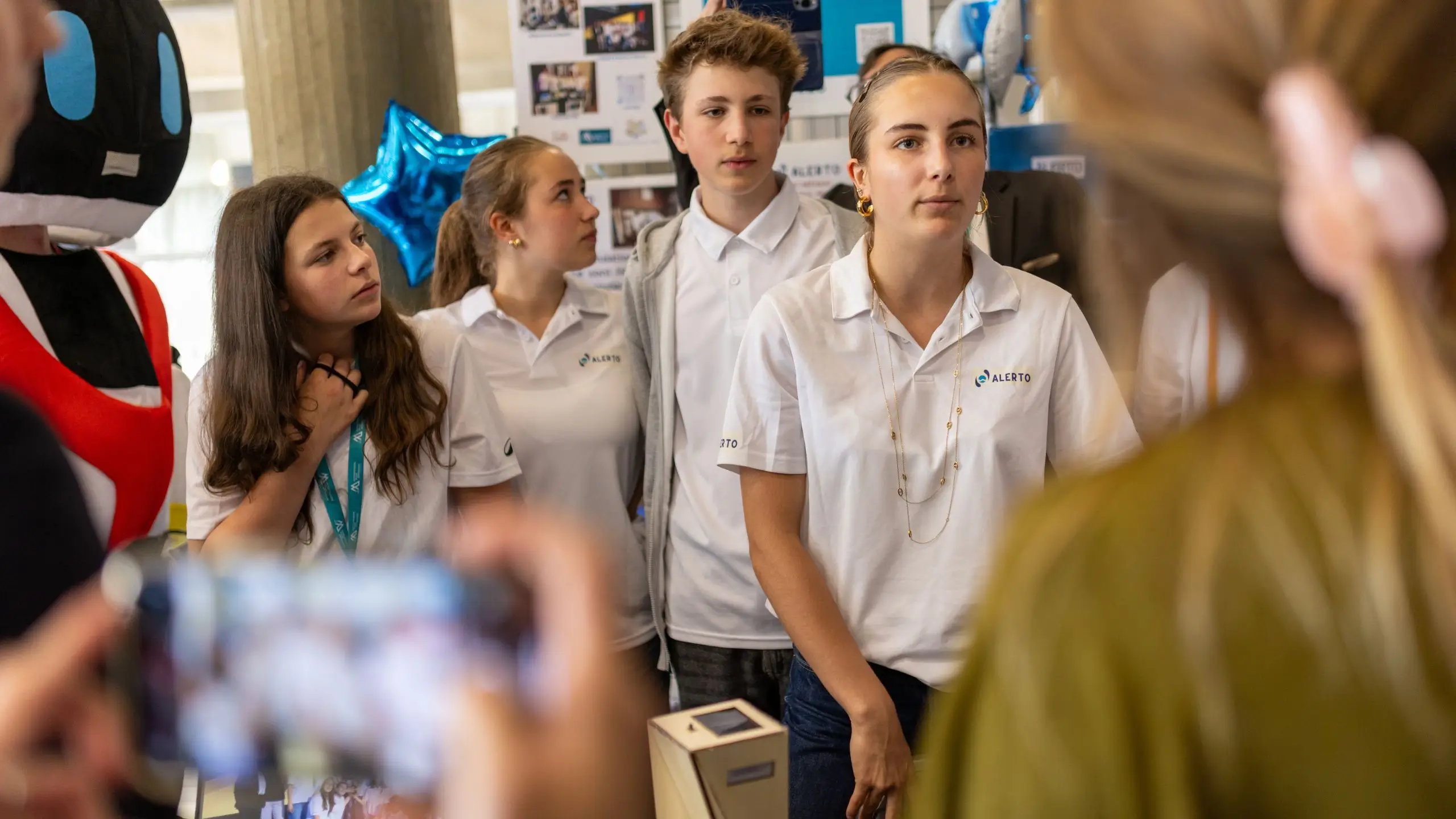 Groupe de jeunes en polos blancs Alerto présentant un projet lors d'un événement, avec une personne floutée prenant une photo au premier plan.