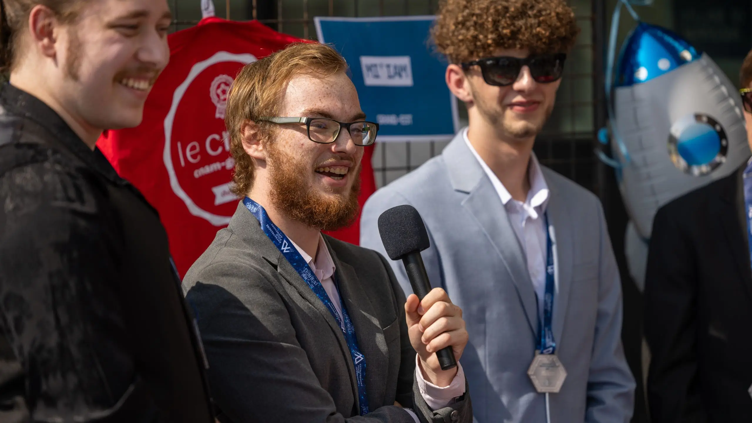 Un jeune homme avec une barbe et des lunettes souriant et tenant un microphone, accompagné de deux autres jeunes hommes portant des badges et des vestes, lors d'un événement extérieur.
