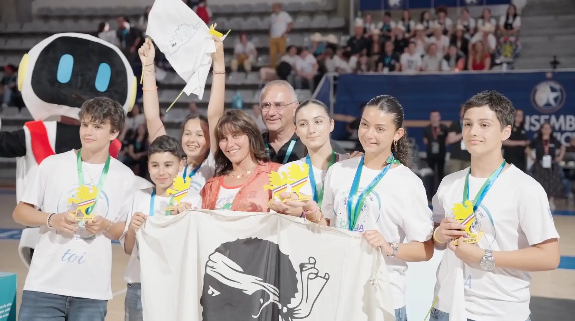Un groupe de jeunes souriants tenant des trophées et une grande bannière lors d'un événement en intérieur avec un public en arrière-plan.