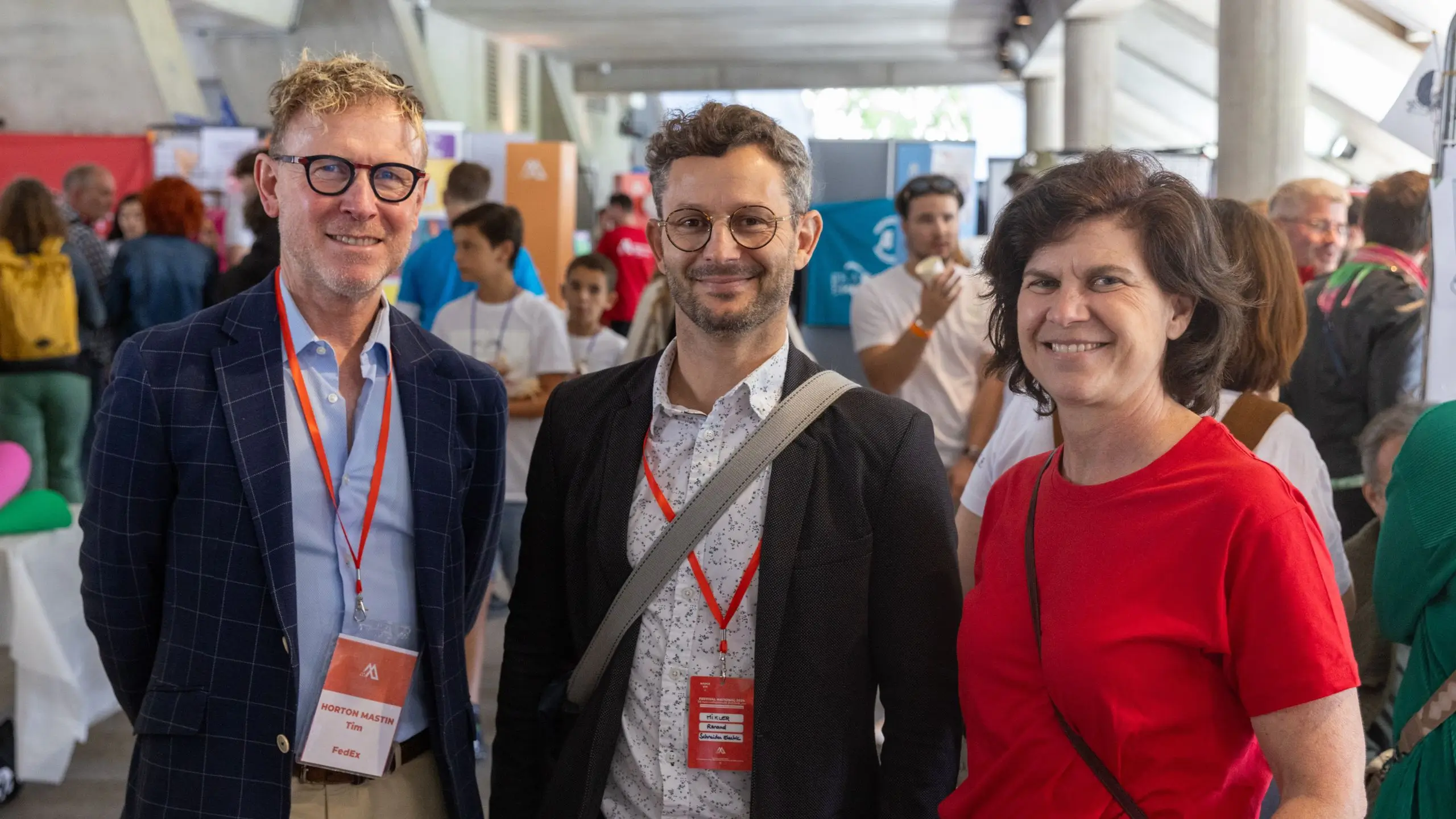 Trois personnes souriantes portant des badges rouges lors d'un événement en intérieur avec foule en arrière-plan.