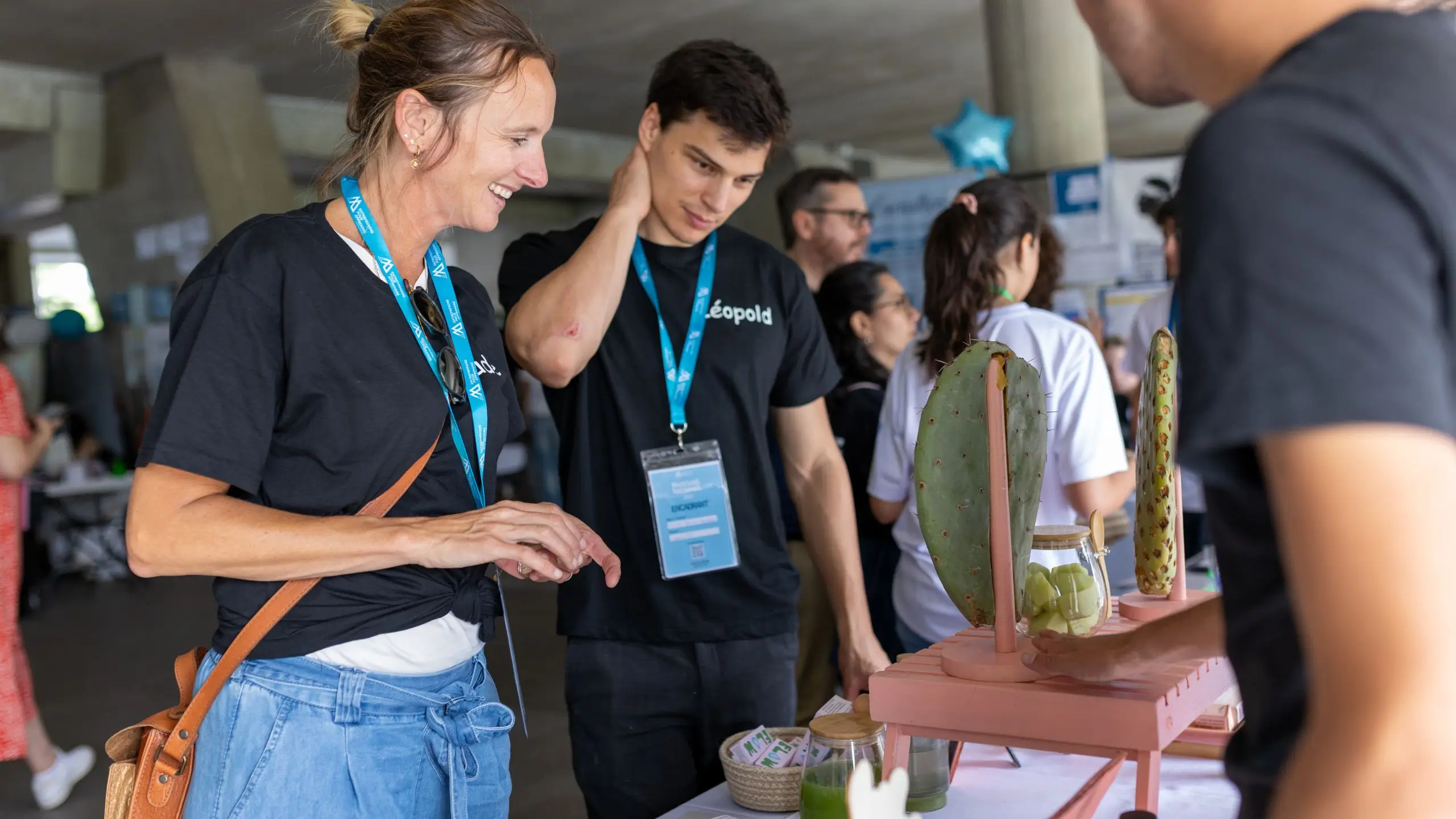 Deux bénévoles souriants discutent à un stand avec des objets présentés sur une table lors d'un événement animé.