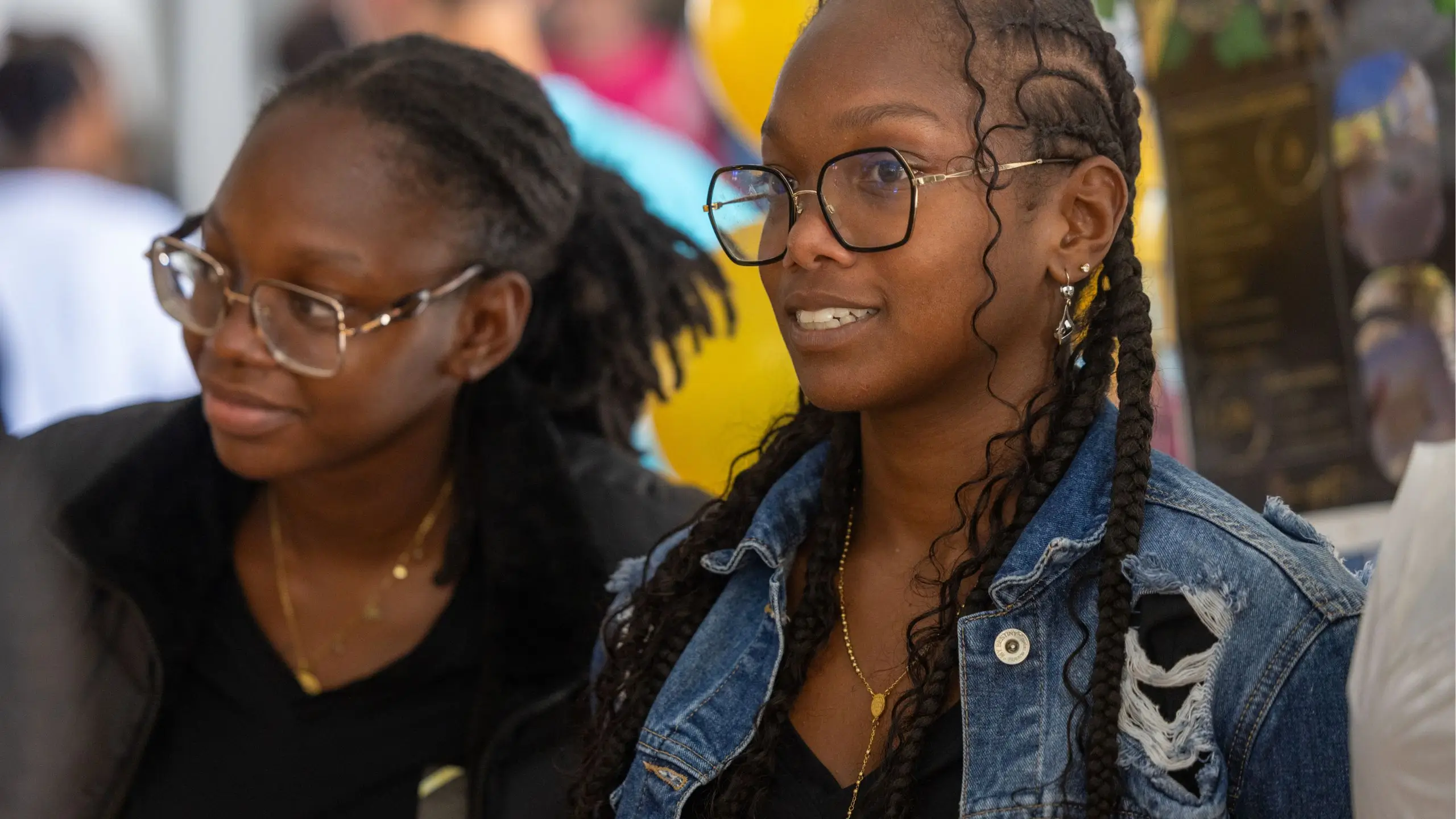 Deux femmes noires portant des lunettes, l'une avec une veste en jean déchirée et des tresses, souriant lors d'un événement.
