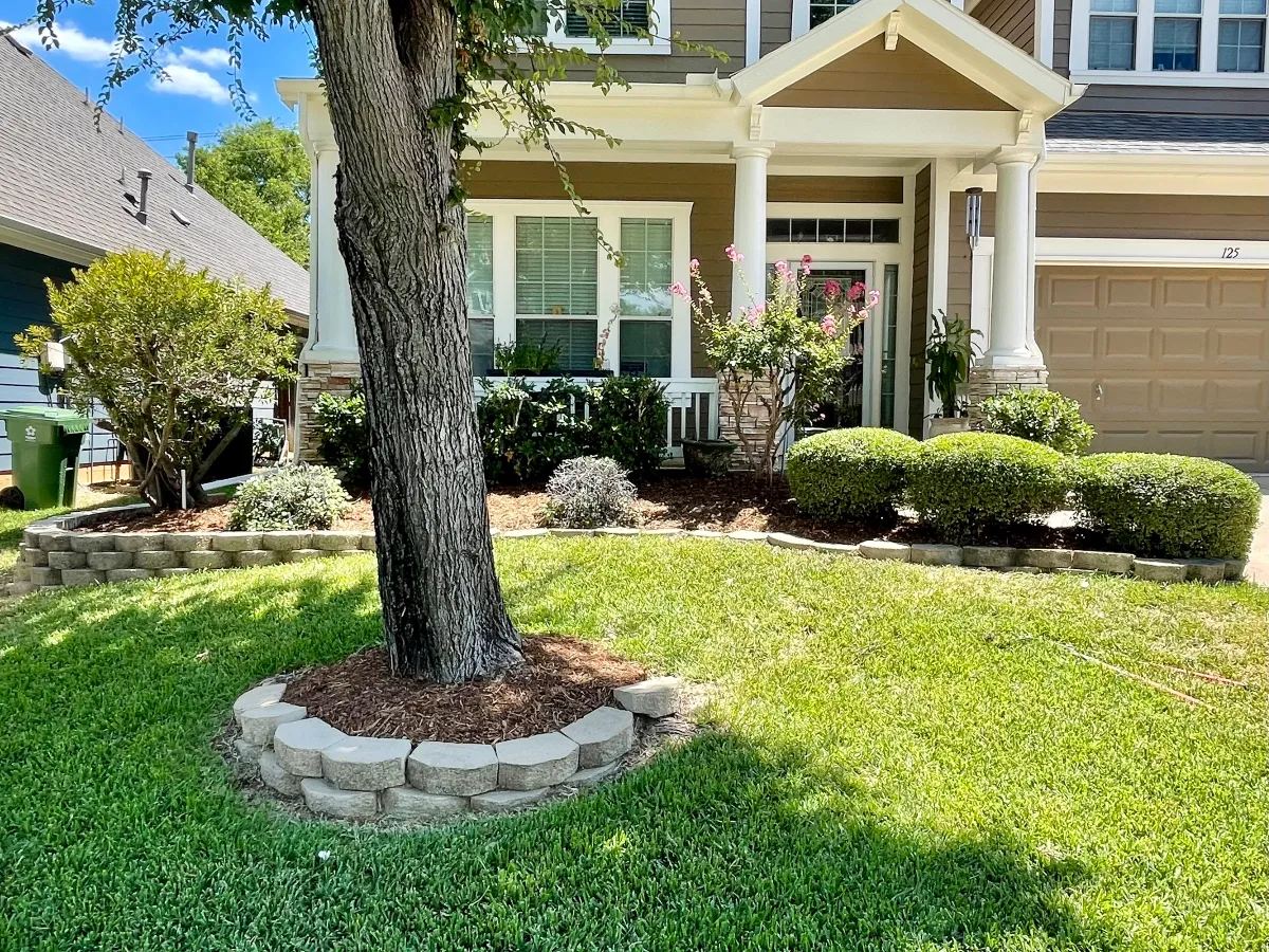 Maintained front yard with mature tree, green lawn, shrubs, and landscaped flower beds