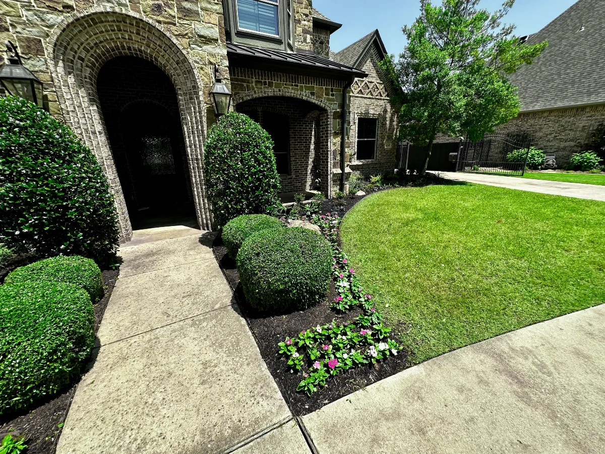 Landscaped stone home entrance with manicured lawn, shrubs, and flowering plants
