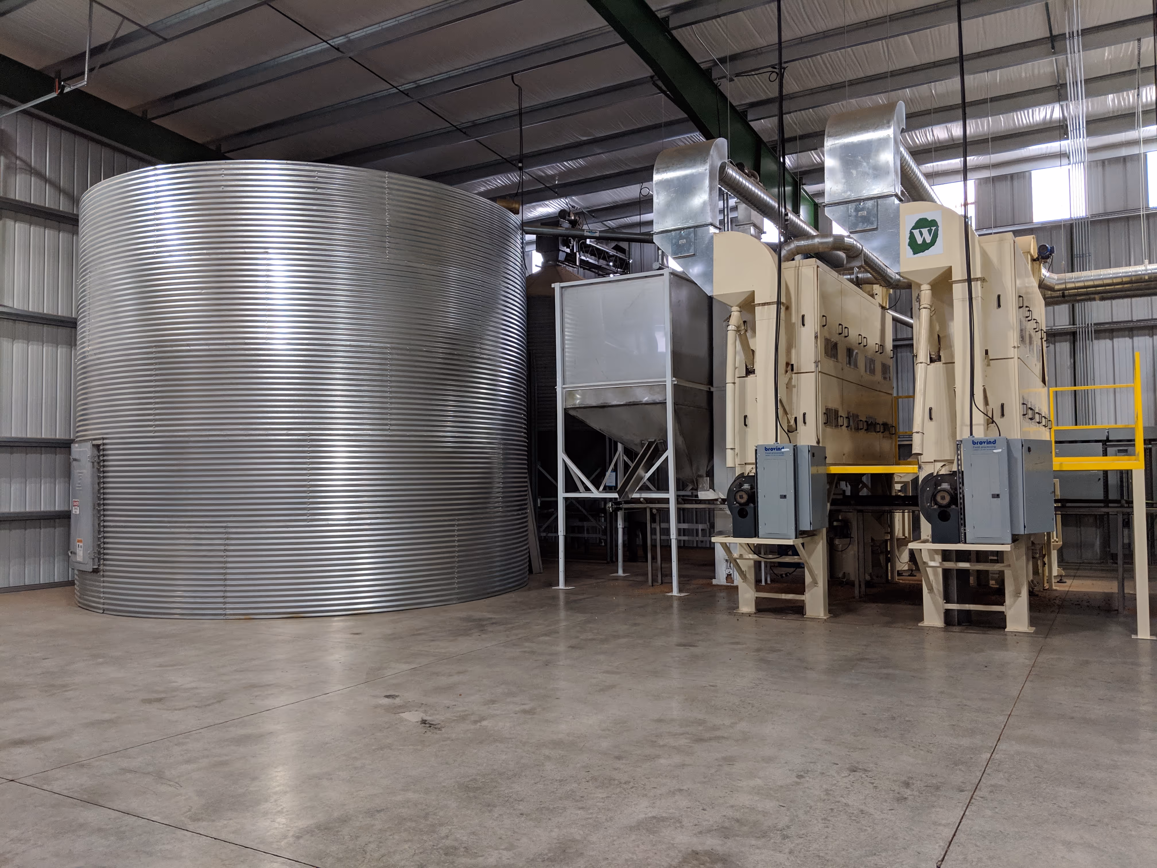 Large industrial metal storage tank next to beige processing machinery inside a spacious warehouse.